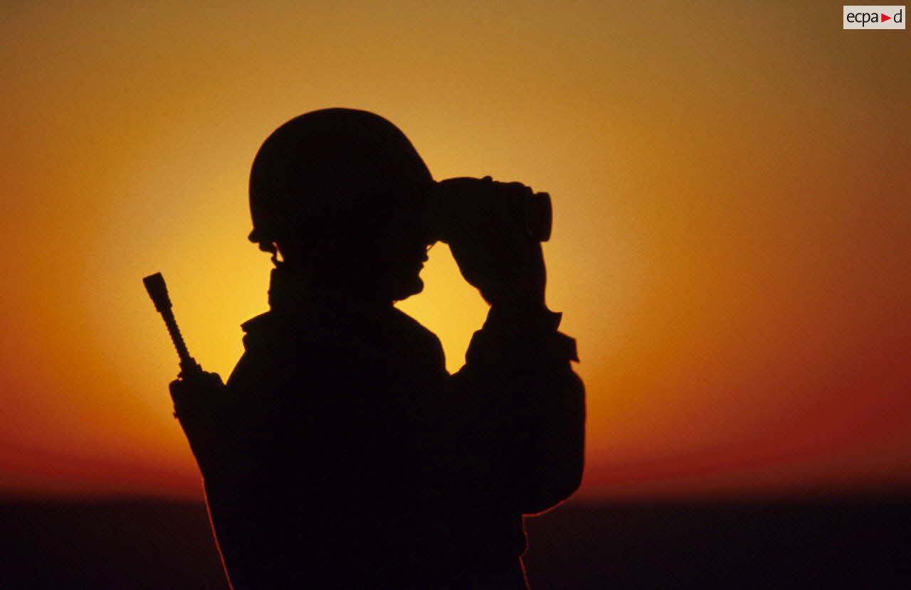En ZDO (zone de déploiement opérationnel) Olive, un soldat est posté en observation dans le désert, équipé de jumelles, en contre-jour au coucher de soleil.