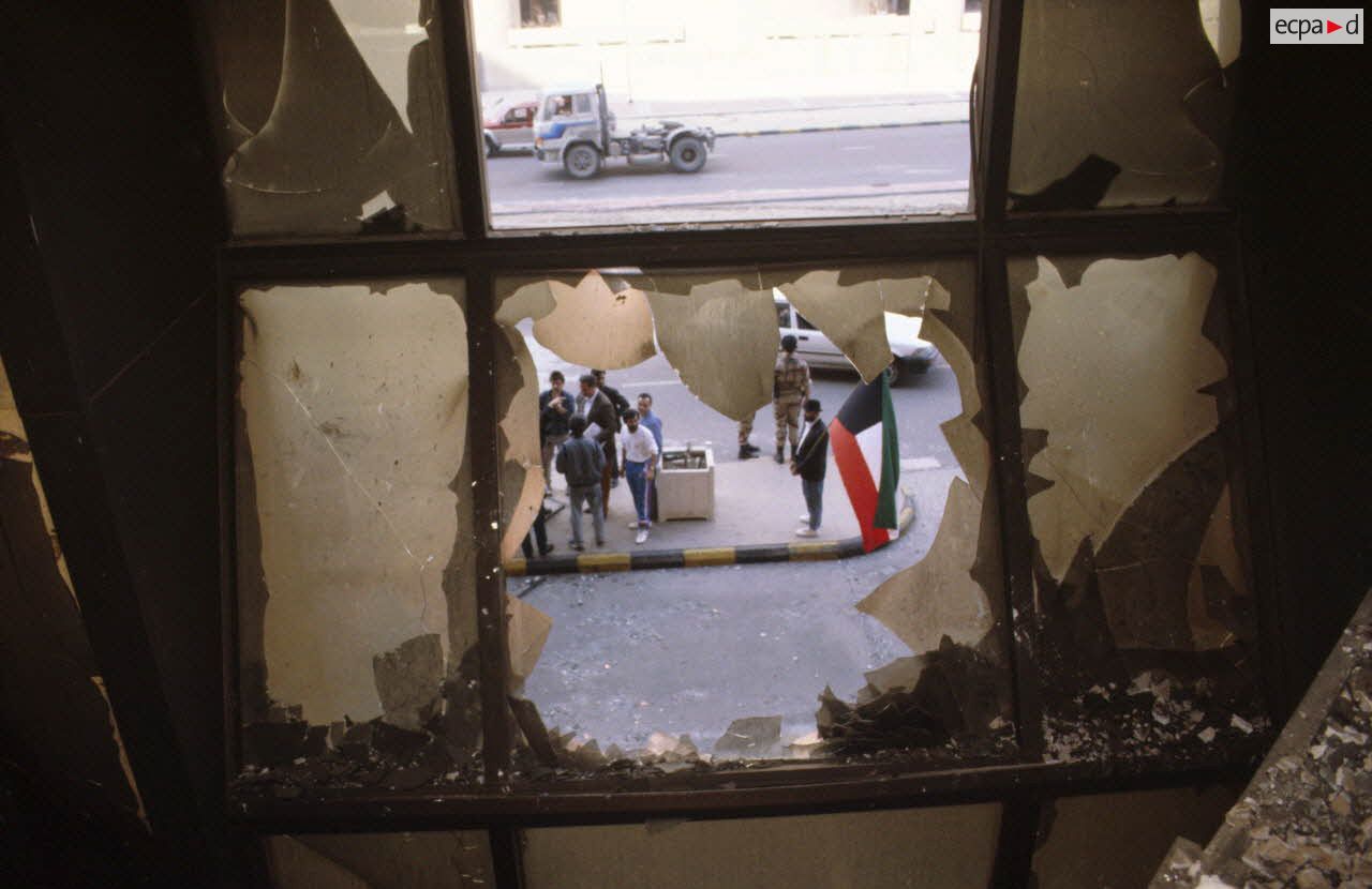 Des civils koweitiens dans la rue vus par une fen&ecirc;tre cass&eacute;e depuis l'int&eacute;rieur carbonis&eacute; de l'h&ocirc;tel Sheraton d&eacute;truit par les militaires irakiens.