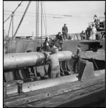 Chargement, à l'aide d'un palan, de torpilles dans les tubes lance-torpilles d'une tourelle orientable d'un sous-marin, à quai dans un port.