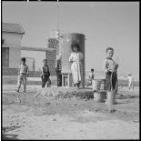 Centre de regroupement de M'Cid. Des enfants à la fontaine sur la place, derrière eux l'école.