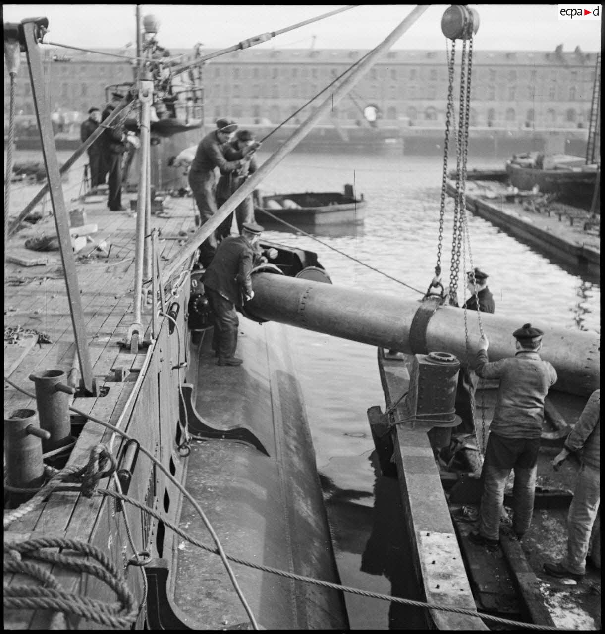 Chargement de torpilles, à l'aide d'un palan, dans les tubes lance-torpilles d'une tourelle orientable d'un sous-marin, à quai dans un port.