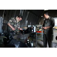 Un sergent-chef armurier ("petaf") sur Mirage 2000 examine des pièces mécaniques sur un établi sous un hangar de maintenance, en présence d'un mécanicien qatari.