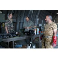 Un sergent-chef armurier ("petaf") sur Mirage 2000, examinant des pièces mécaniques sur un établi sous un hangar de maintenance, en présence de deux mécaniciens qataris.