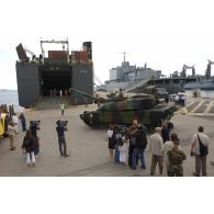 Le char Leclerc Bayeux du 6/12e RC (régiment de cuirassiers), à l'embarquement pour le Liban, monte la rampe d'accès du cargo affrété Fast Arrow sur le port de Toulon, guidé par un personnel du 601e RCR (régiment de circulation routière), en présence de caméramen et de photographes. En arrière-plan, le BAP (bâtiment-atelier polyvalent) de la Marine nationale Jules Verne à quai.