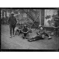 [Des soldats sont assis et allongés sur le bord d'une route de village, pendant que l'un d'entre eux répare la roue d'un vélo.]