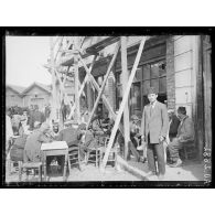 [Scène de la vie quotidienne : des hommes sont réunis autour de tables devant un batiment en construction.]