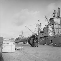 Activités en zone portuaire de Moruroa, le bâtiment de soutien santé (BSS) Rance (A618) et le bâtiment de soutien logistique (BSL) Tarn (A771) à quai. Un engin de manutention transporte un rouleau de câble de transmission. Sur un container est inscrit "Ballon".