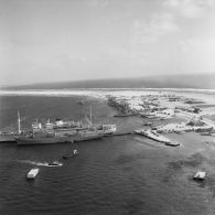 Vue aérienne de la zone portuaire de Moruroa, de bâtiments à quai, de la rade et de la force Alfa au mouillage. En arrière-plan, la piste d'atterrissage.