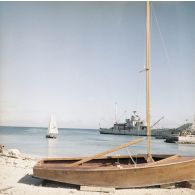 Au port de Moruroa, derrière un petit dériveur sur cales, deux personnels naviguent à voile près du rivage, non loin de la gabare Scorpion (A728) à quai.