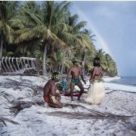 Un couple de danseurs, coiffés d'une couronne fleurie, fait une démonstration de danse du Tamoure, accompagnés par un guitariste sur une plage de l'atoll d'Hao. En arrière-plan, un arc-en-ciel.