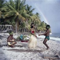 Un couple de danseurs, coiffés d'une couronne fleurie, fait une démonstration de danse du Tamoure, accompagnés par un guitariste sur une plage de l'atoll d'Hao. En arrière-plan, un arc-en-ciel.