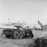 Sur un quai du port de Moruroa, un légionnaire manoeuvre un engin tracteur devant un camion chargé sur un chaland. En arrière-plan, le bâtiment-base Maurienne (A637) à quai.