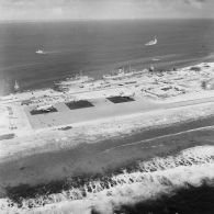 Vue aérienne de la zone aéroportuaire de l'atoll de Moruroa. Avions Breguet Deux Ponts au parking de l'aéroport, et en arrière-plan, le bâtiment de soutien santé (BSS) Rance (A618) et le bâtiment de soutien logistique (BSL) Tarn (A771) à quai au port. Des bâtiments de la force Aplha dans le lagon, dont peut-être le TCD Ouragan.