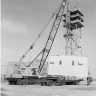 Levage d'un shelter au-dessus d'une remorque par un camion grue P&H sur le site du poste de commande de tir (PCT) Anémone au pied d'une antenne de transmission à Moruroa.