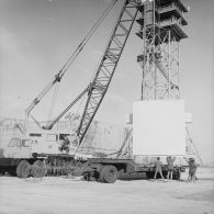 Levage d'un shelter au-dessus d'une remorque par un camion grue P&H sur le site du poste de commande de tir (PCT) Anémone au pied d'une antenne de transmission à Moruroa.