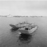 Vue aérienne de chalands de transport de matériel (CTM) chargés de camions dans le lagon de Moruroa. En arrière-plan, le bâtiment-base Médoc (A612) à quai.