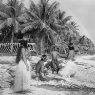 Un couple de danseurs, coiffés d'une couronne fleurie, fait une démonstration de danse du Tamoure, accompagnés par un guitariste sur une plage de l'atoll d'Hao. Inscription "Hao" sur une épave d'embarcation en arrière-plan.