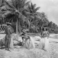 Un couple de danseurs, coiffés d'une couronne fleurie, a fait une démonstration de danse du Tamoure, accompagnés par un guitariste sur une plage de l'atoll d'Hao. Inscription "Hao" sur une épave d'embarcation en arrière-plan.