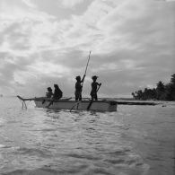 Sur la plage de Hao, groupe d'habitants en tenue traditionnelle à bord d'une pirogue de pêcheurs à balancier, dont une jeune femme jouant de la guitare.