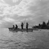 Sur la plage de Hao, groupe d'habitants en tenue traditionnelle à bord d'une pirogue de pêcheurs à balancier, dont une jeune femme jouant de la guitare.