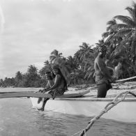 Sur la plage de Hao, groupe d'habitants en tenue traditionnelle à bord d'une pirogue de pêcheurs à balancier, dont une jeune femme jouant de la guitare.