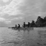 Sur la plage de Hao, groupe d'habitants en tenue traditionnelle à bord d'une pirogue de pêcheurs à balancier, dont une jeune femme jouant de la guitare.