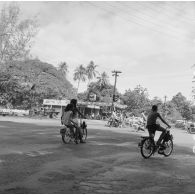 Circulation des deux-roues, vélos et solex, dans une rue qui croise le cours de l'Union sacrée, à Papeete, non loin d'une station service.