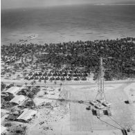 Vue aérienne d'une antenne du site du poste de commande de tir (PCT) Anémone sur l'atoll de Moruroa, devant une zone vie avec des fare de Moruroa. Au bout de la jetée, un bâtiment-atelier (peut-être L9082).