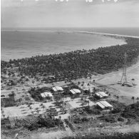 Vue aérienne d'une antenne du site du poste de commande de tir (PCT) Anémone sur l'atoll de Moruroa, devant une zone vie avec des fare de Moruroa. En arrière-plan, des bâtiments de la force Alpha au mouillage et les bâtiments-bases au port.