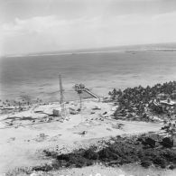 Vue aérienne du site du poste de commande de tir (PCT) Anémone sur l'atoll de Moruroa. Au bout de la jetée, deux bâtiments-ateliers (dont peut-être L9082). Un bâtiment la force Alpha dans le lagon. En arrière-plan, le port avec des bâtiments-bases à quai.