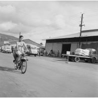 Camions au chargement de marchandises devant un hangar du port de Papeete. Un couple circule en solex.