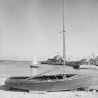 Au port de Moruroa, derrière un petit dériveur sur cales, deux personnels naviguent à voile près du rivage, non loin de la gabare Scorpion (A728) à quai.