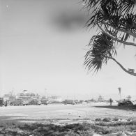 Au port de l'atoll de Moruroa, une flotte de véhicules Deux-chevaux Citroën sur un quai. En arrière-plan, le bâtiment-base (BB) Maurienne (A637) et le bâtiment de soutien santé (BSS) Rance (A618).