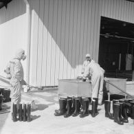 Devant un hangar du service de décontamination de Hao, des personnels en tenue de protection lavent des bottes dans un bac après un tir d'essai nucléaire.