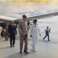 Le général Charles De Gaulle, président de la République, et le vice-amiral Jean Lorain, commandement du groupe opérationnel des expérimentations nucléaires (GOEN), suivis de Pierre Messmer, ministre des Armées, au pied de l'avion à l'aéroport de Moruroa. En arrière-plan, Pierre Billotte, ministre des Départements et territoires d'Outre-mer, et Jean Viard, directeur des essais.