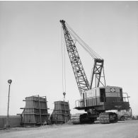 Construction de la digue du port de Papeete. Un engin chenillé à grue Bucyrus-Erie lève un coffrage à béton de talus de digue.