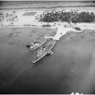 Vue aérienne d'un site portuaire de Moruroa avec l'aviso-colonial Francis Garnier (F730) et le landing craft tank ou utility (LCT ou LCU) L9088 ou L9089 à quai et un chantier à l'arrière-plan sur l'atoll. [vue inversée]