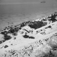 Vue aérienne d'installations sur l'atoll. Hangars. L'aviso-colonial Francis Garnier F730 au mouillage. Peut-être l'atoll de Moruroa.