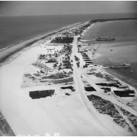 Vue aérienne d'installations du site sur l'atoll. Zone de stockage, barils d'essence, silos, aviso-colonial Francis Garnier F730 et landing craft tank (LCT) L9098 à quai. Peut-être l'atoll de Moruroa.