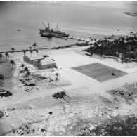 Vue aérienne d'installations sur l'atoll. Aviso-colonial Francis Garnier F730 et navire-atelier annexe L9099 (ancien landing craft tank ou LCT) à quai. Zone de poser d'hélicoptères Alouette II. Peut-être l'atoll de Moruroa.