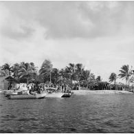 Vue depuis l'eau du rivage et du camp militaire. Zone de stockage de barils, canot à moteur, zodiac et ponton. Peut-être la base Hortensia sur l'atoll de Moruroa.