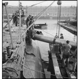 Chargement de torpilles, à l'aide d'un palan, dans les tubes lance-torpilles d'une tourelle orientable d'un sous-marin, à quai dans un port.