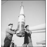 Deux marins manipulent un obus de 330 mm suspendu à un palan à bord du cuirassé Dunkerque.