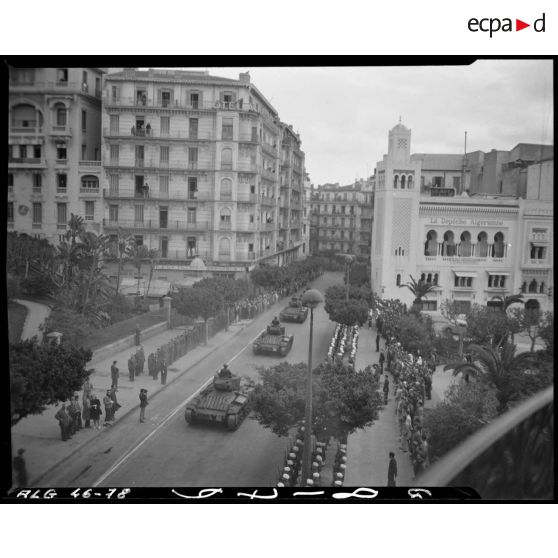 Plan en plongée d'un défilé motorisé, sous le regard d'autorités militaires et civiles.