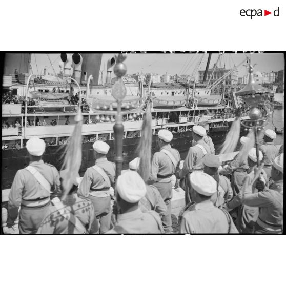 Plan rapproché de l'arrivée du bateau de jeunes gens originaires de Saint-Etienne par la musique d'une unité de l'armée d'Afrique, au port d'Alger.