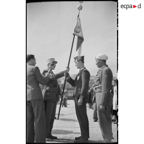 Une personnalité militaire remet un drapeau à un jeune garçon originaire de Saint-Etienne, sous le regard d'un journaliste qui enregistre l'évènement, sur le port d'Alger.