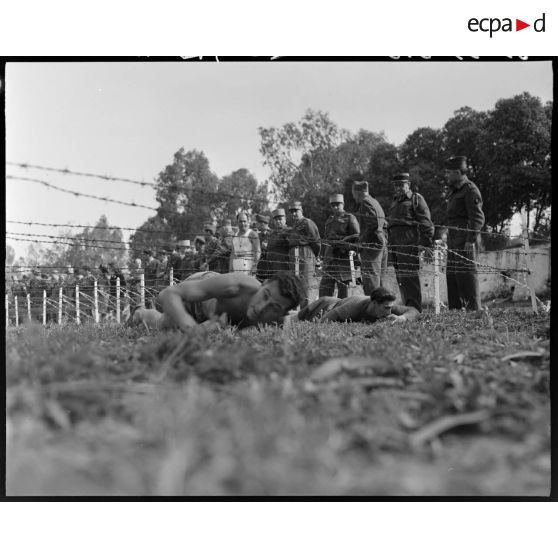 Des aides moniteurs d'éducation physique militaire (EPM) rampent sous des barbelés bas, sous le regard d'autorités militaires.