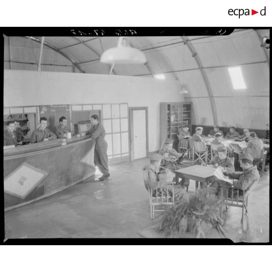 Plan en plongée d'un espace commun du camp du Lido, à l'intérieur duquel des soldats lisent autour de tables et se désaltèrent au comptoir d'un bar.