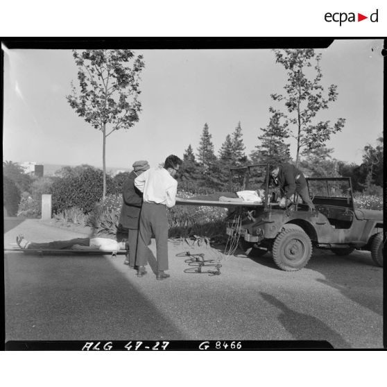 Des hommes fixent deux brancards sur lesquels sont allongées deux personnes, à l'arrière d'une voiture Jeep transformée en transport sanitaire.