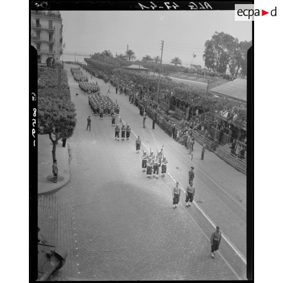 Plan en plongée d'une unité militaire qui défile sous le regard d'autorités civiles et militaires.